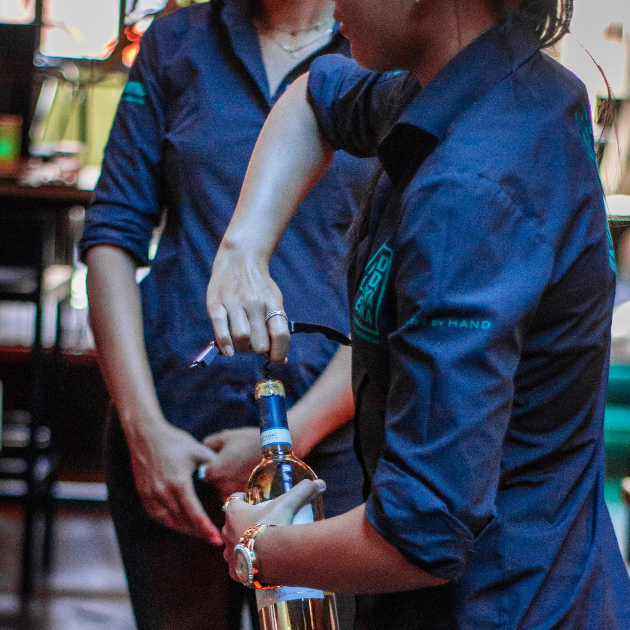 Two women wearing uniforms while bartending. Elevate your staff with custom uniforms from Stamford Uniform and Linen.