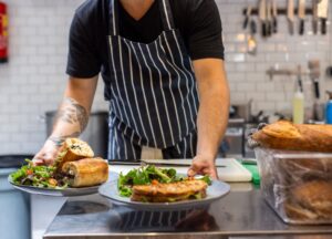 A chef in a striped apron serves plated sandwiches and Linen.