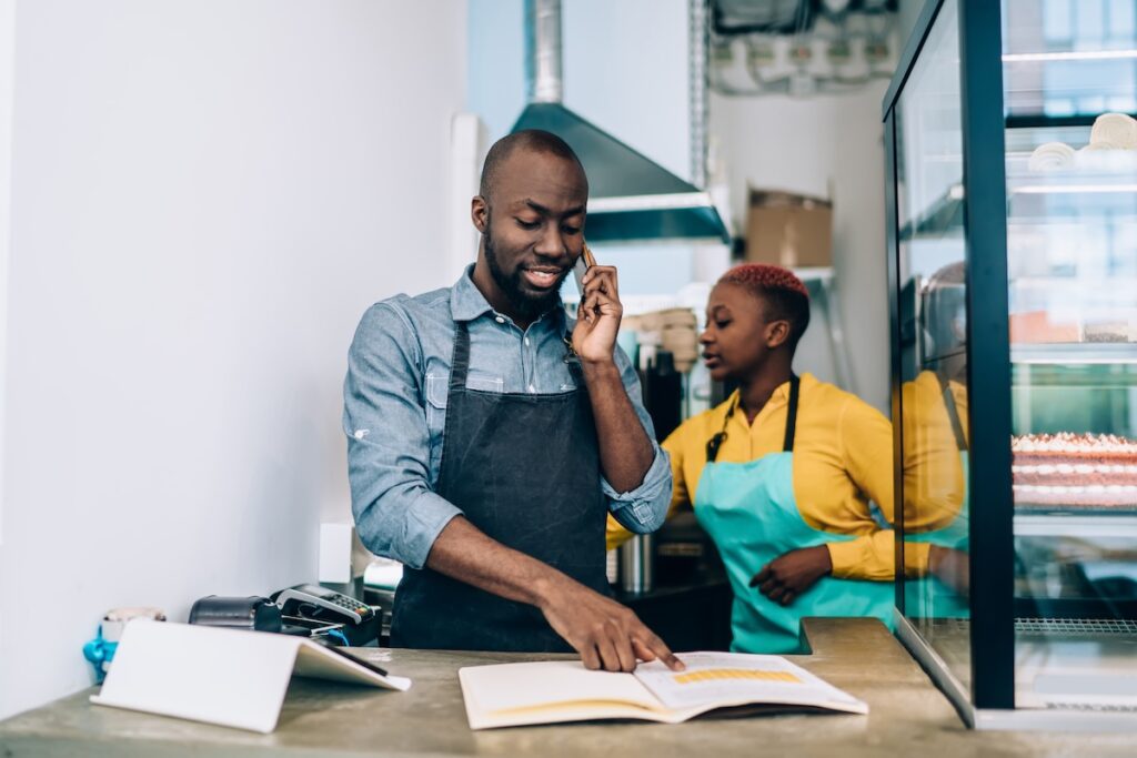 African American male in apron checking information in open notepad while standing behind counter and having smartphone conversation in small restaurant
